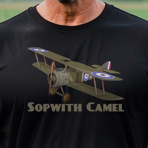 Puede incluir: Una camiseta negra con un gráfico de un avión Sopwith Camel. El avión es marrón y verde con detalles en rojo, blanco y azul. El texto "Sopwith Camel" está impreso debajo del avión.