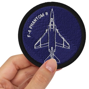 May include: A blue and black embroidered patch with a white outline of an F-4 Phantom II jet fighter. The text "F-4 PHANTOM II" is embroidered in white around the edge of the patch.