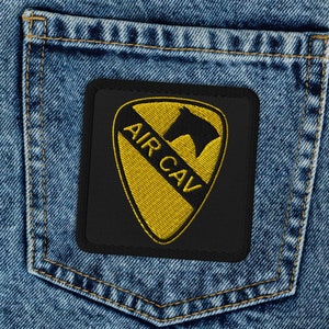 Puede incluir: Un parche negro con un galón amarillo y una silueta de caballo negro. El parche dice "AIR CAV" en letras amarillas. El parche está cosido en un bolsillo de mezclilla azul.