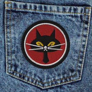 May include: Blue denim jeans with a red and black patch featuring a stylized black cat with yellow eyes on the back pocket.