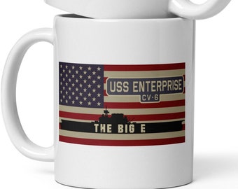 Pode incluir: Duas canecas de cerâmica branca empilhadas uma sobre a outra. Cada caneca apresenta o texto "USS ENTERPRISE CV-6" e "THE BIG E" sobre um desenho que se assemelha à bandeira americana. As canecas têm um acabamento brilhante e uma alça curva.