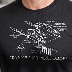 May include: Black t-shirt featuring a detailed white diagram of a missile launcher, including labels like "Terrier Missile Round" and "MK 5 MOD 3 GUIDED MISSILE LAUNCHER." The shirt is worn by a man with gray hair and a beard.