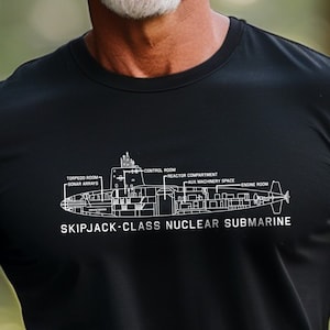 Può includere: Una maglietta nera con un'illustrazione bianca di un sottomarino nucleare di classe Skipjack. L'illustrazione mostra i diversi compartimenti del sottomarino, tra cui la sala siluri, le antenne sonar, la sala di controllo, il compartimento del reattore, la sala macchine e lo spazio macchine di poppa.
