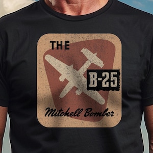 May include: A black t-shirt with a vintage-style graphic of a B-25 Mitchell Bomber. The graphic is in a red, white, and brown color scheme and features the text "THE B-25 Mitchell Bomber".