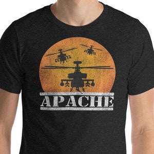 May include: A black t-shirt featuring a vintage-style graphic of three Apache helicopters flying in front of a sunset. The text "APACHE" is printed below the helicopters.