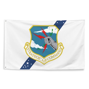 May include: White flag featuring the Strategic Air Command emblem. The emblem includes a shield with a blue background, a silver armored arm holding red lightning bolts, and the text "STRATEGIC AIR COMMAND". A blue diagonal stripe with white stars is also present.