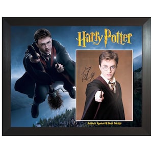 Puede incluir: Un marco que muestra el logotipo de Harry Potter, una imagen de un personaje volando en una escoba y un retrato de Daniel Radcliffe sosteniendo una varita con una firma auténtica. El marco es de color marrón oscuro y rectangular.
