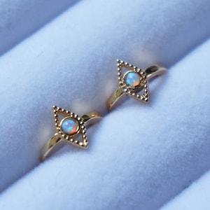 May include: Two gold rings with a white opal gemstone in the center of each ring. The rings are in the shape of a diamond with a beaded border.