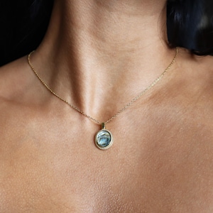 May include: A gold-toned necklace featuring a circular pendant with a textured, beaded edge. The centre of the pendant contains an iridescent, swirling abalone shell inlay with blue, green, and grey tones, suspended from a delicate gold-toned chain.