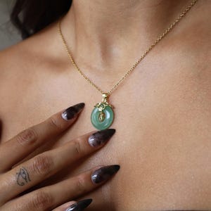 Könnte beinhalten: Goldfarbene Halskette mit Jade-Anhänger. Der Anhänger ist hellgrün und rund, mit einer goldenen Verzierung. Die Halskette wird um den Hals getragen.