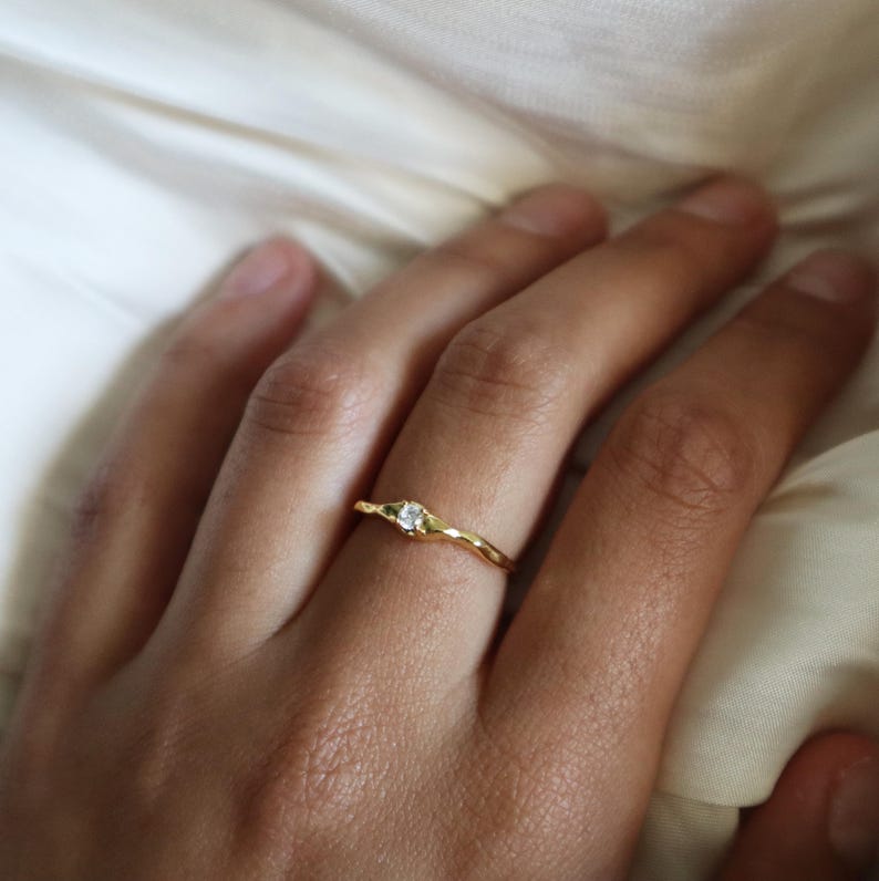 May include: A gold ring with a single, clear gemstone set in the center. The ring has a textured, hammered finish.