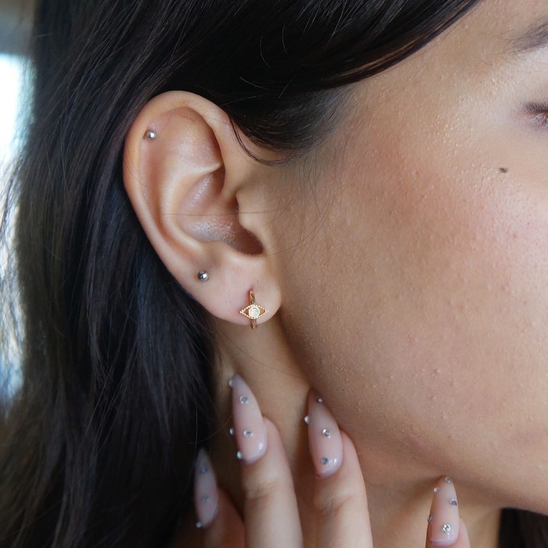 May include: A close-up of a person's ear with two small earrings. The earring on the lower lobe is a gold hoop with a small white opal stone in the shape of an eye. The earring on the upper lobe is a small silver stud.