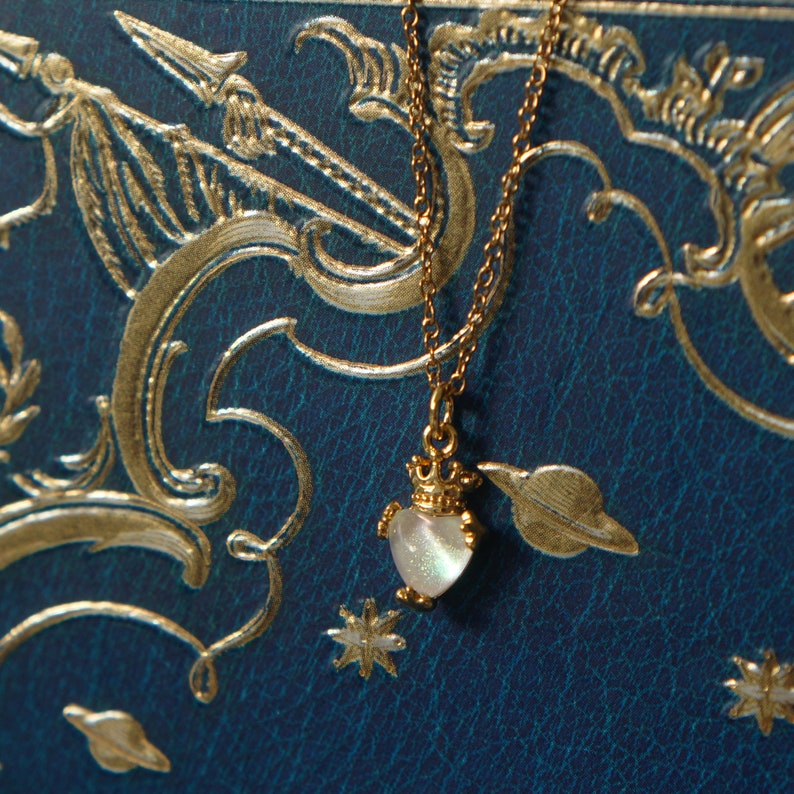 May include: A gold chain necklace with a small, iridescent heart pendant. The pendant has a crown on top and is set against a blue background with gold accents.