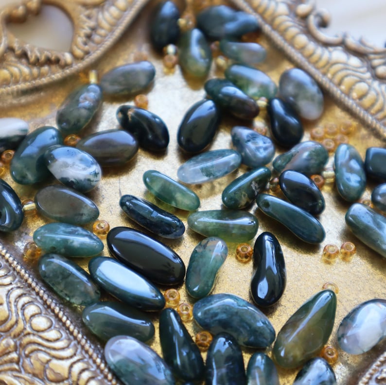 May include: A collection of polished, oval-shaped moss agate stones in varying shades of green and black. The stones are scattered on a decorative, gold-colored tray with ornate edges. The stones have a smooth, glossy finish.