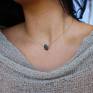 May include: A delicate gold necklace featuring a teardrop-shaped, dark green pendant. The pendant hangs from a thin gold chain, creating a minimalist and elegant design. The necklace is shown on a person wearing a light-colored, open-weave top.
