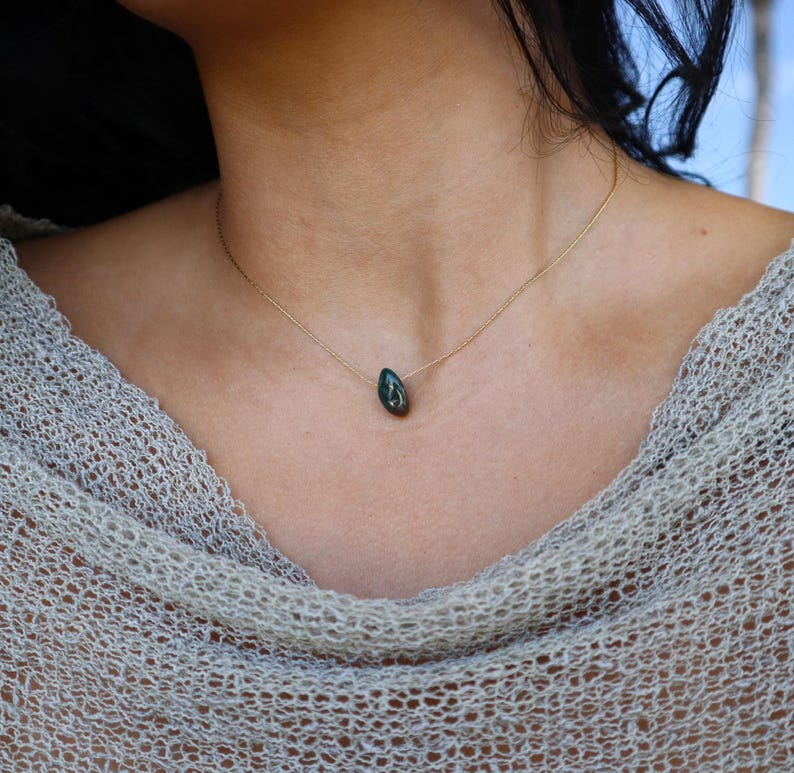 May include: A delicate gold necklace featuring a teardrop-shaped, dark green pendant. The pendant hangs from a thin gold chain, creating a minimalist and elegant design. The necklace is shown on a person wearing a light-colored, open-weave top.