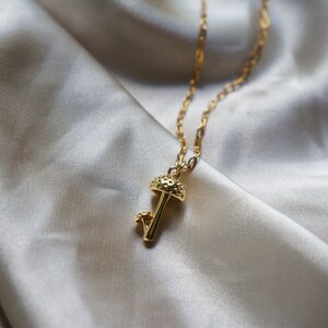 Puede incluir: Un collar dorado con un colgante de hongo. El colgante presenta un dise&ntilde;o detallado de hongo con un sombrero texturizado y un hongo m&aacute;s peque&ntilde;o al lado. La cadena dorada es delicada y est&aacute; sobre un fondo satinado.