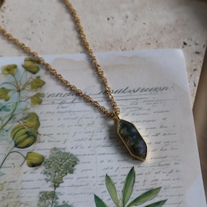 Könnte beinhalten: Eine goldfarbene Halskette mit einem Anhänger aus einem dunkelblauen und grünen Stein. Der Anhänger hat einen goldfarbenen Rahmen und eine einzigartige, unregelmäßige Form. Die Kette hat ein filigranes, gedrehtes Design. Die Halskette liegt auf einem Vintage-Papierhintergrund.