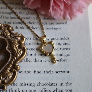 May include: A gold necklace with a heart-shaped pendant. The pendant is made of mother-of-pearl and has a keyhole design. The necklace is on a book with text.