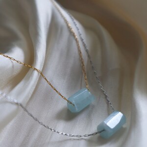 May include: Two delicate necklaces with light blue gemstone pendants. The necklaces are layered on a white fabric background. The top necklace has a gold chain and the bottom necklace has a silver chain.