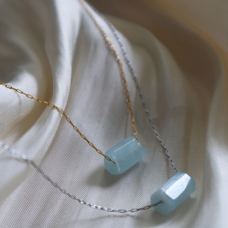 May include: Two delicate necklaces with light blue gemstone pendants. The necklaces are layered on a white fabric background. The top necklace has a gold chain and the bottom necklace has a silver chain.