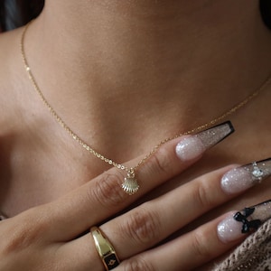 May include: A gold chain necklace with a small seashell pendant. The necklace is worn by a person with long, glittery fingernails.