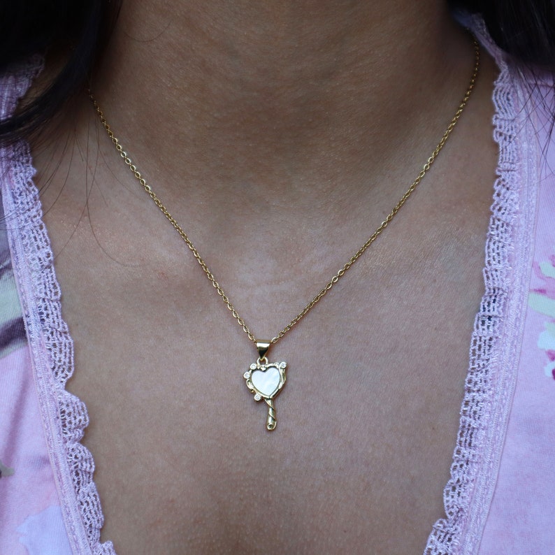 May include: A gold chain necklace with a heart-shaped pendant. The pendant is a mirror with a white background and a gold frame. The frame is decorated with small, clear stones.