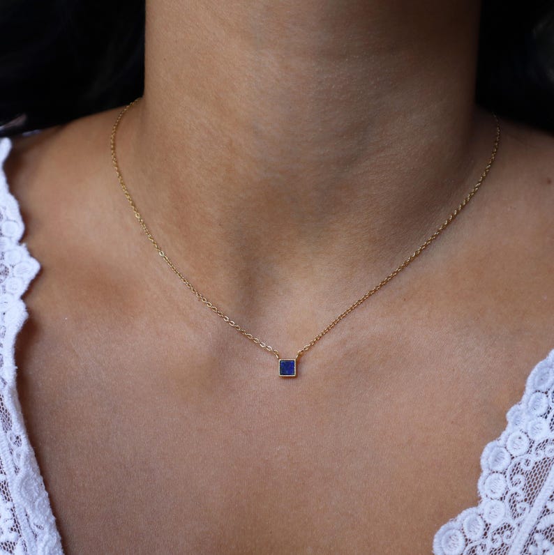 Peut inclure: Un collier d&eacute;licat en cha&icirc;ne dor&eacute;e avec un petit pendentif de forme carr&eacute;e orn&eacute; d'une pierre pr&eacute;cieuse bleu fonc&eacute;.