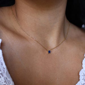 Peut inclure: Un collier d&eacute;licat en cha&icirc;ne dor&eacute;e avec un petit pendentif en forme de carr&eacute; de pierre pr&eacute;cieuse bleue.