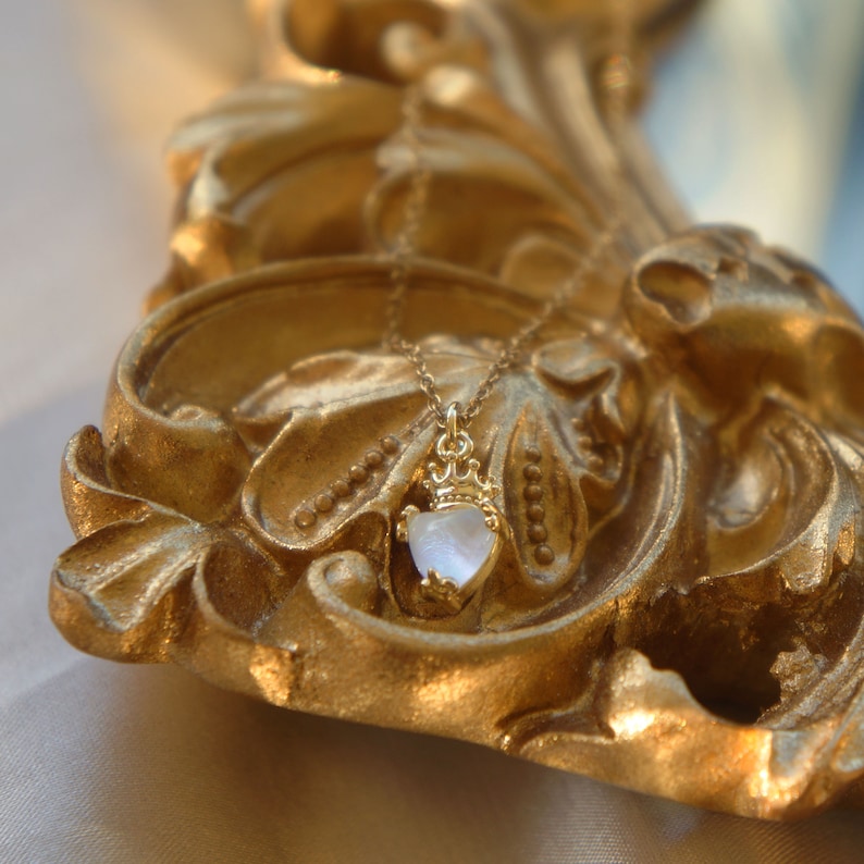 May include: A gold necklace with a heart-shaped pendant featuring a white iridescent stone. The necklace is displayed on a gold ornate frame.