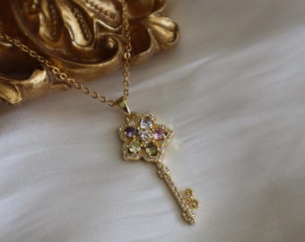 May include: A gold key necklace with a flower-shaped charm. The charm is adorned with colorful gemstones and clear rhinestones.