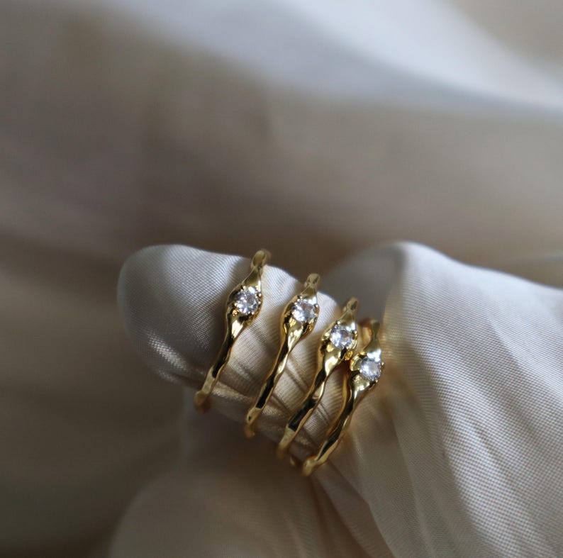 May include: Four gold rings with a single clear stone set in each. The rings are stacked on top of each other and are on a white fabric background.
