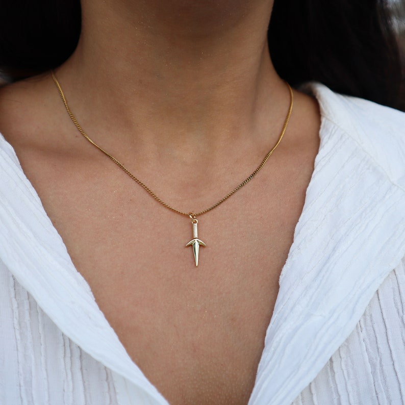Puede incluir: Un collar de cadena dorada con un peque&ntilde;o colgante de daga. El collar lo lleva una persona con una camisa blanca.