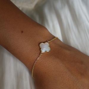 May include: A gold-colored bracelet featuring a four-leaf clover charm with a white, iridescent center. The bracelet is delicate and appears to be made of linked chain. The charm is set in a gold-colored frame. The bracelet is worn on a wrist.