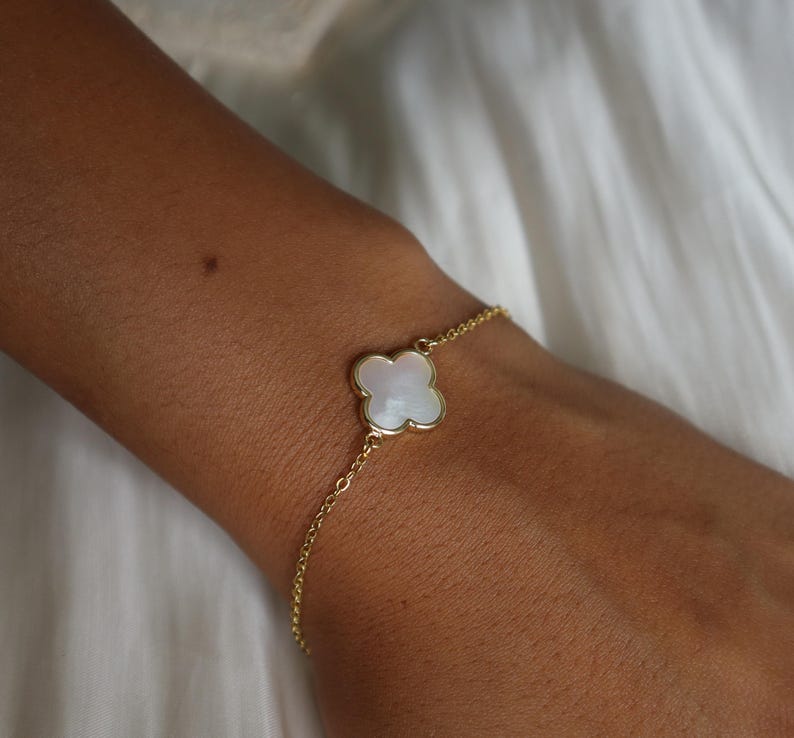 May include: A gold-colored bracelet featuring a four-leaf clover charm with a white, iridescent center. The bracelet is delicate and appears to be made of linked chain. The charm is set in a gold-colored frame. The bracelet is worn on a wrist.