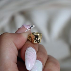 May include: A gold-colored charm in the shape of a small cup with a textured surface.