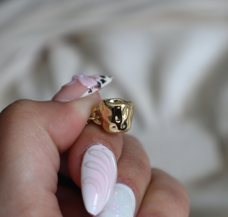 May include: A gold-colored charm in the shape of a small cup with a textured surface.
