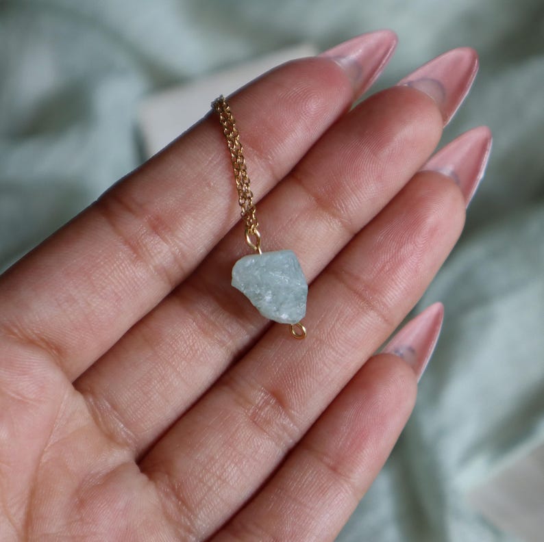 May include: A gold-colored chain necklace with a small, light blue, raw aquamarine stone pendant. The stone is irregularly shaped and has a rough texture. The necklace is displayed against a soft, blurred background.