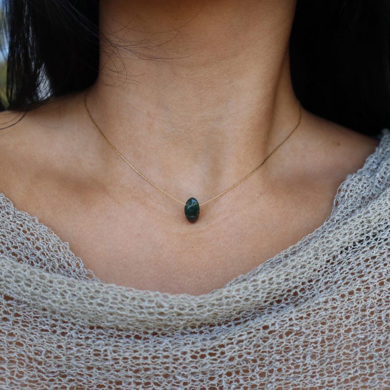 May include: A delicate gold necklace featuring a dark green, oval-shaped gemstone pendant. The pendant is suspended from a thin gold chain, creating a minimalist and elegant design. The necklace is shown against a neutral background.