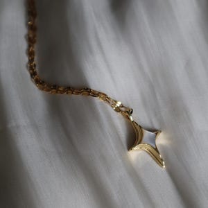May include: A gold-toned necklace with a starburst pendant. The pendant features a white, pearlescent center framed by a gold border. The chain is a delicate, linked design. The necklace is displayed on a white, textured surface.
