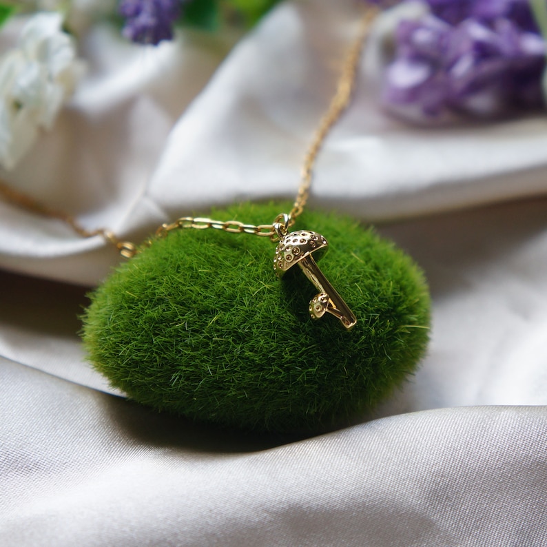 Puede incluir: Un collar con colgante de hongo dorado. El colgante presenta un sombrero y un tallo texturizados, suspendidos de una fina cadena. El collar descansa sobre un lecho de musgo verde, sobre un fondo de tela satinada.