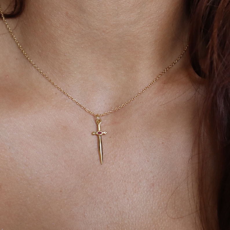 May include: A gold-colored pendant necklace featuring a small sword charm with a red gemstone. The sword charm is suspended from a delicate chain. The necklace is worn around the neck.
