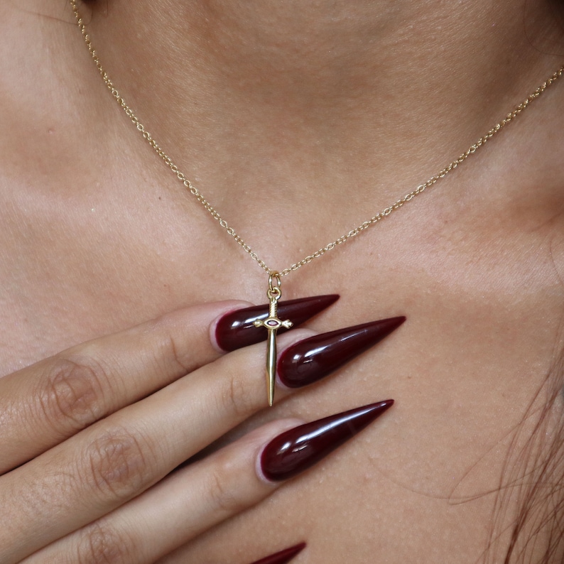 May include: A gold-colored sword pendant necklace. The pendant has a small red and white accent. The necklace is displayed on a person's skin. The nails are painted a dark red color.