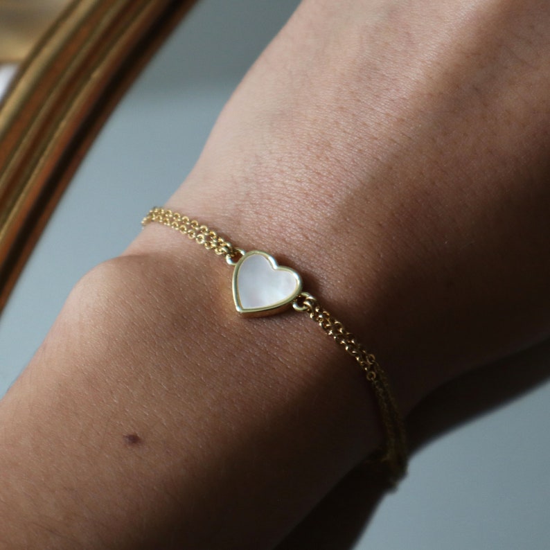 May include: A gold chain bracelet with a white heart-shaped charm. The charm has a slightly iridescent finish.