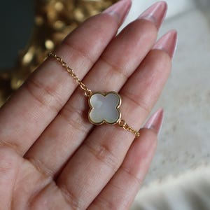 May include: Gold-toned chain bracelet featuring a four-leaf clover charm. The charm has a white, pearlescent center and a gold-colored border. The bracelet is delicate and appears to be made of metal.