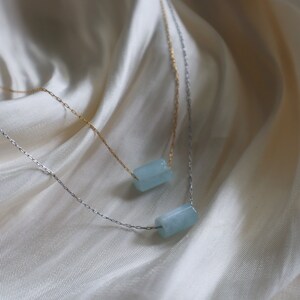 May include: Two delicate necklaces with light blue rectangular stones hanging on a white fabric background.
