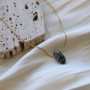 May include: A delicate gold-colored chain necklace featuring a dark gray, oval-shaped gemstone pendant. The necklace is displayed on a white, silky fabric, with a textured, light-colored stone in the background.