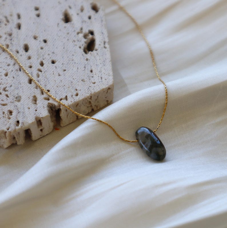 May include: A delicate gold-colored chain necklace featuring a dark gray, oval-shaped gemstone pendant. The necklace is displayed on a white, silky fabric, with a textured, light-colored stone in the background.