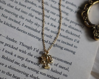 May include: A gold-colored necklace with a pendant shaped like a set of scales. The necklace is on a chain and is displayed on a page of a book with printed text. The scales pendant is detailed and has a small base.