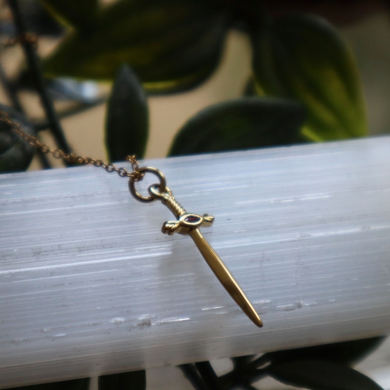 May include: A gold-colored pendant necklace featuring a miniature sword design. The sword has a detailed hilt and a pointed blade. The necklace chain is delicate and also gold-toned. The background is a blurred view of green foliage and a white, textured surface.